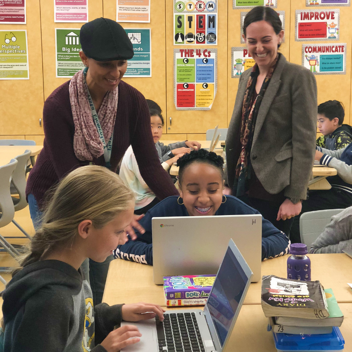 Two female teachers with two students in a classroom looking at laptops